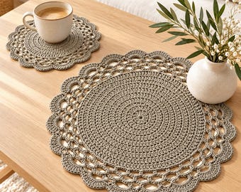 May include: Two round, taupe-colored crocheted mats featuring a dense, solid center and a decorative, scalloped openwork border. One mat supports a ceramic mug filled with coffee, while the other holds a white vase with green foliage.