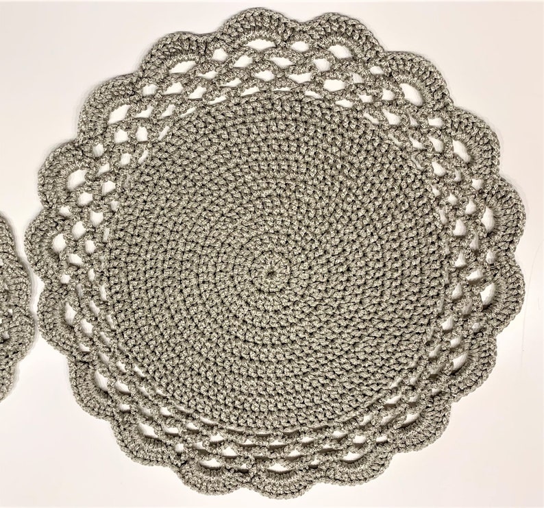 May include: Two gray crocheted placemats. The placemats are round with a scalloped edge. The center of the placemats have a spiral design. The placemats are made of a soft, textured yarn.