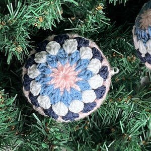 May include: Handmade crochet Christmas ornament with a floral design. The ornament features a pink center, surrounded by white, light blue, and navy blue crochet petals. The ornament is hanging on a green Christmas tree.