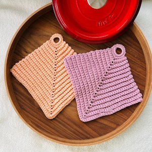 Könnte beinhalten: Zwei handgefertigte Topflappen aus Häkelarbeit in Pfirsich und Lavendel, mit einer Schlaufe zum Aufhängen. Die Topflappen sind quadratisch und liegen auf einem Holztablett neben einem roten Kochtopf.