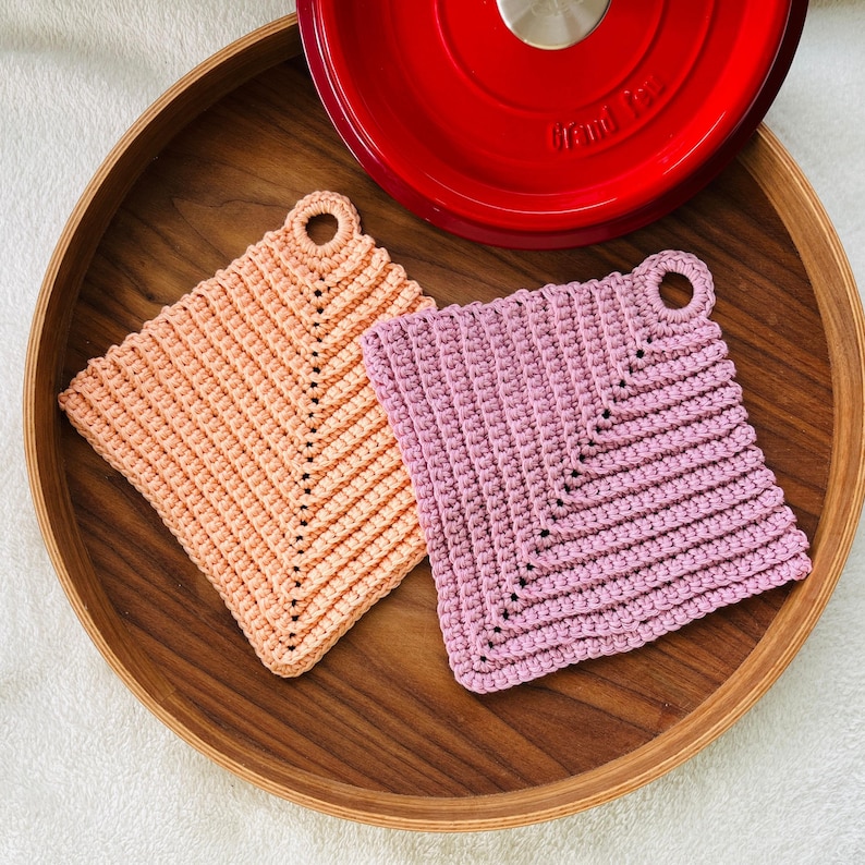 Op de afbeelding: Twee handgemaakte gehaakte pannenlappen in perzik en lavendel, met een lus om op te hangen. De pannenlappen zijn vierkant en liggen op een houten dienblad, naast een rode kookpot.