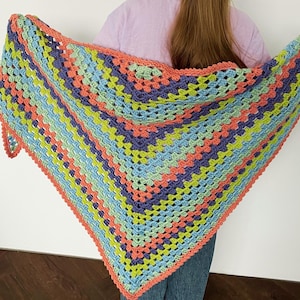 May include: A colorful, handmade crochet shawl in a triangular shape. The shawl features a vibrant mix of colors, including blue, green, purple, and coral. The edges are finished with a coral border, creating a visually appealing and cozy accessory.