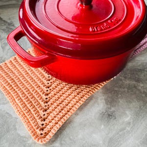 Op de afbeelding: Een felrode ge&euml;mailleerde gietijzeren braadpan met bijpassende deksel, rustend op een perzikkleurige gehaakte pannenlap. De pot heeft een donkere houten handgreep op het deksel en twee zijhandgrepen. De pannenlap is vierkant en heeft een textuurpatroon.