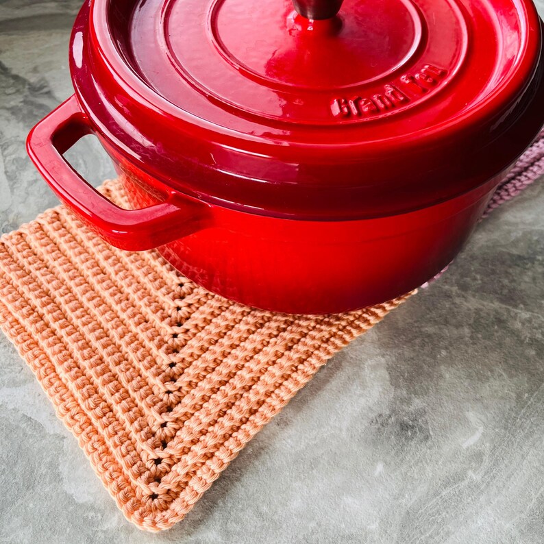 Op de afbeelding: Een felrode ge&euml;mailleerde gietijzeren braadpan met bijpassende deksel, rustend op een perzikkleurige gehaakte pannenlap. De pot heeft een donkere houten handgreep op het deksel en twee zijhandgrepen. De pannenlap is vierkant en heeft een textuurpatroon.