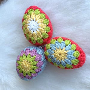 Three crocheted Easter eggs in different colors and patterns. The eggs are red, blue, and pink with yellow and green accents.
