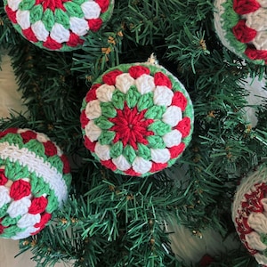 Pode incluir: Enfeites de Natal feitos &agrave; m&atilde;o em croch&ecirc; vermelho, verde e branco. Os enfeites redondos apresentam um padr&atilde;o detalhado e colorido. S&atilde;o exibidos em galhos verdes de &aacute;rvore de Natal, criando uma cena festiva.