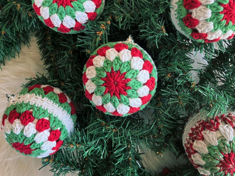 Pode incluir: Enfeites de Natal feitos &agrave; m&atilde;o em croch&ecirc; vermelho, verde e branco. Os enfeites redondos apresentam um padr&atilde;o detalhado e colorido. S&atilde;o exibidos em galhos verdes de &aacute;rvore de Natal, criando uma cena festiva.