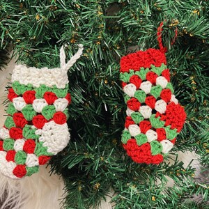 Pode incluir: Duas meias de Natal de croch&ecirc; feitas &agrave; m&atilde;o. As meias s&atilde;o vermelhas, brancas e verdes, com punho e la&ccedil;o para pendurar brancos. S&atilde;o exibidas em um fundo de &aacute;rvore de Natal verde.