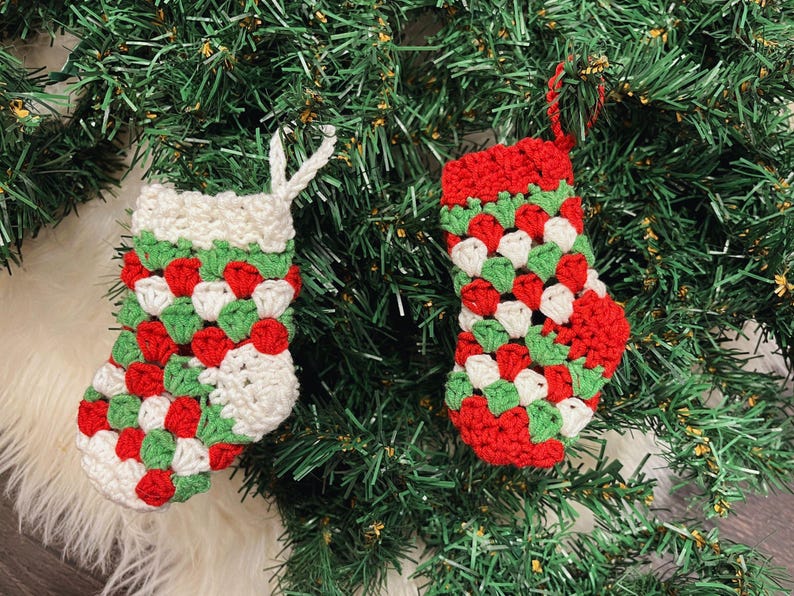 Pode incluir: Duas meias de Natal de croch&ecirc; feitas &agrave; m&atilde;o. As meias s&atilde;o vermelhas, brancas e verdes, com punho e la&ccedil;o para pendurar brancos. S&atilde;o exibidas em um fundo de &aacute;rvore de Natal verde.