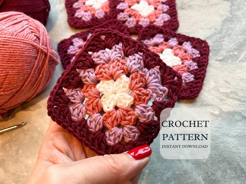 May include: Close-up of a crocheted granny square in shades of burgundy, pink, and cream. The square is held in a hand, with yarn and a crochet hook visible in the background. Text on the image reads "CROCHET PATTERN INSTANT DOWNLOAD."