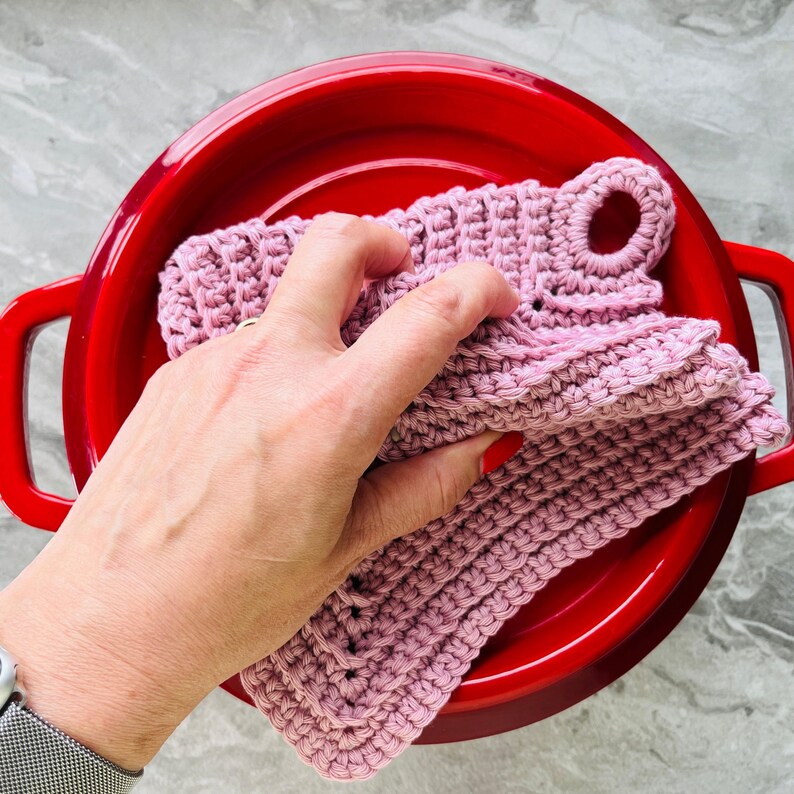 Op de afbeelding: Een hand houdt een stapel roze gehaakte pannenlappen vast met een lus om op te hangen. De pannenlappen worden in een rode ge&euml;mailleerde kookpot met handvatten geplaatst. De pannenlappen zijn gemaakt van zacht, getextureerd garen.