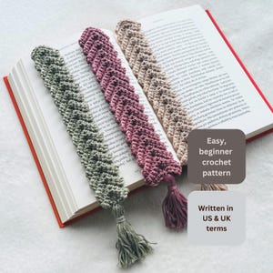 Può includere: Tre segnalibri all'uncinetto fatti a mano nei colori verde salvia, rosa antico e beige, ciascuno caratterizzato da un motivo a festoni testurizzato e da una nappa in filo coordinata. I segnalibri sono appoggiati sulle pagine di un libro aperto.