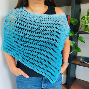 Puede incluir: Un poncho de ganchillo azul, con un degradado de claro a turquesa. El diseño de tejido abierto crea una sensación ligera y aireada. El poncho se lleva sobre una camiseta negra y vaqueros azules, ofreciendo un look casual y elegante. Mide aproximadamente 100 cm de largo.