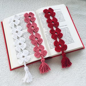 May include: Three handmade crochet flower bookmarks in white, pink, and red, each with a matching tassel. The bookmarks are placed on the pages of an open book with red covers, showcasing the intricate floral design.