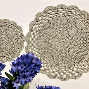 May include: Two crocheted placemats in a neutral gray color. The larger mat has a scalloped edge, while the smaller one is round. The mats are made with a spiral design. A bouquet of blue flowers is in the foreground.