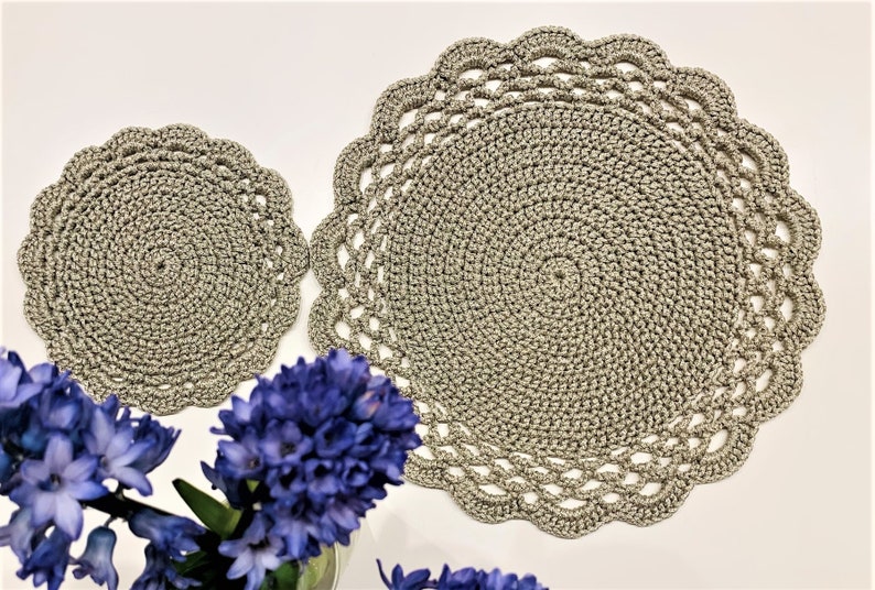 May include: Two crocheted placemats in a neutral gray color. The larger mat has a scalloped edge, while the smaller one is round. The mats are made with a spiral design. A bouquet of blue flowers is in the foreground.