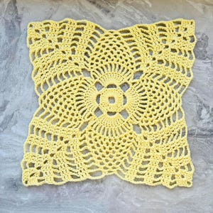 May include: A square, pale yellow crochet doily featuring an intricate, openwork pattern with a central circular motif surrounded by radiating shell-like stitches and a delicate, lacy border that creates a geometric, four-pointed star shape.