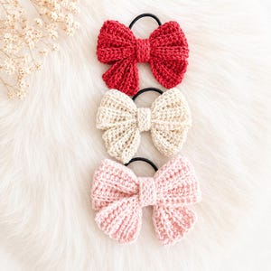 May include: Three hair ties featuring crocheted bows in red, cream, and light pink. Each bow is constructed with a ribbed texture and attached to a simple black elastic band, providing a secure way to hold hair in place with a soft, textile finish.