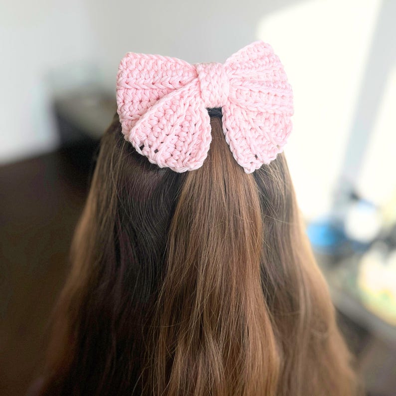 May include: A light pink crochet hair bow accessory featuring a textured, ribbed stitch pattern. The bow is constructed with two large, symmetrical loops and a central knot detail, designed to be secured to hair for a decorative finish.