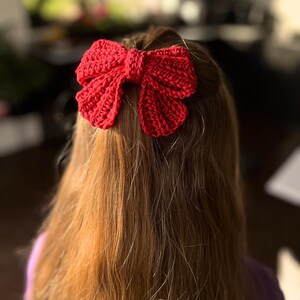 May include: A bright red hair accessory shaped like a bow, crafted with a textured crochet stitch pattern. The bow features a central knot detail and two distinct loops on each side, creating a voluminous appearance when secured in long hair.