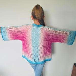 May include: A handmade, open-weave crochet shrug in a gradient of pink, light blue, and white. The sleeves and neckline are trimmed with a darker blue. The garment is lightweight and appears to be made of soft yarn.