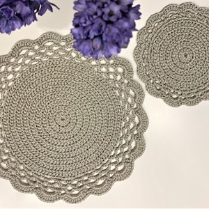 May include: Two gray crocheted placemats and a matching coaster are displayed on a white surface, with purple flowers in the background. The placemats have a scalloped edge and a spiral design. The coaster is round with a similar design.