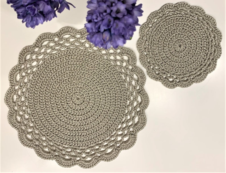 May include: Two gray crocheted placemats and a matching coaster are displayed on a white surface, with purple flowers in the background. The placemats have a scalloped edge and a spiral design. The coaster is round with a similar design.