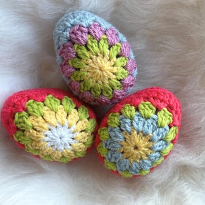 Könnte beinhalten: Drei gehäkelte Ostereier in verschiedenen Farben, darunter rosa, blau, gelb und grün. Die Eier sind mit einem Blumenmuster verziert.