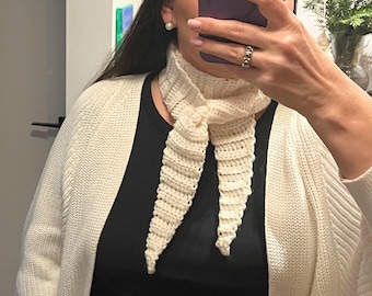 May include: Cream-colored crochet scarf with a pointed end. The scarf is tied in a knot at the neck. The person is wearing a black top and a cream-colored cardigan. The scarf is a handmade accessory.