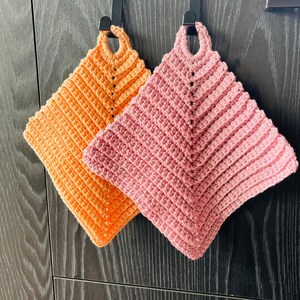 Op de afbeelding: Twee handgemaakte gehaakte pannenlappen in tinten oranje en roze. De pannenlappen zijn vierkant met een lus om op te hangen en een gestructureerd, geribbeld ontwerp. Ze hangen tegen een donkere houten achtergrond.