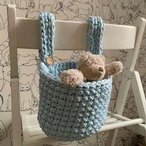 Può includere: Un cestino all'uncinetto fatto a mano, azzurro, con due cinghie, appeso a una sedia bianca. Un piccolo orsacchiotto è all'interno del cestino. Il cestino è fissato con un bottone di legno.