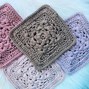 May include: Four handmade crochet squares in shades of pink, brown, and lavender. Each square features a floral design in the centre, surrounded by a textured border. The squares are arranged on a soft, white surface.