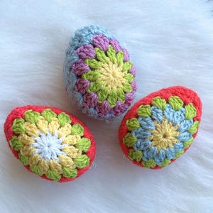 Three crocheted Easter eggs in different colors. The eggs are decorated with a colorful flower pattern. The eggs are on a white fluffy surface.