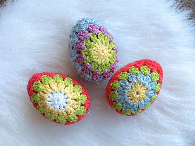 Three crocheted Easter eggs in different colors. The eggs are decorated with a colorful flower pattern. The eggs are on a white fluffy surface.