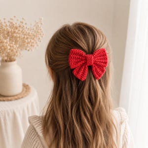 May include: A bright red crocheted hair bow featuring a textured, ribbed stitch pattern. The bow is secured at the center with a matching red yarn band, creating a classic shape that is attached to the back of long, light brown hair.