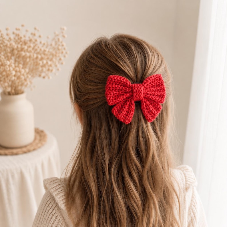 May include: A bright red crocheted hair bow featuring a textured, ribbed stitch pattern. The bow is secured at the center with a matching red yarn band, creating a classic shape that is attached to the back of long, light brown hair.
