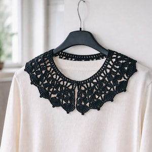 May include: A black, crocheted collar with a scalloped edge is displayed on a white sweater. The collar is intricately designed with openwork patterns and is suspended from a black hanger against a white wall.