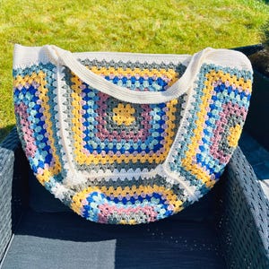 May include: A handmade crochet tote bag with a colorful granny square design. The bag features a cream-colored strap and border, with squares of yellow, blue, pink, and gray. The bag is sitting on a black chair.