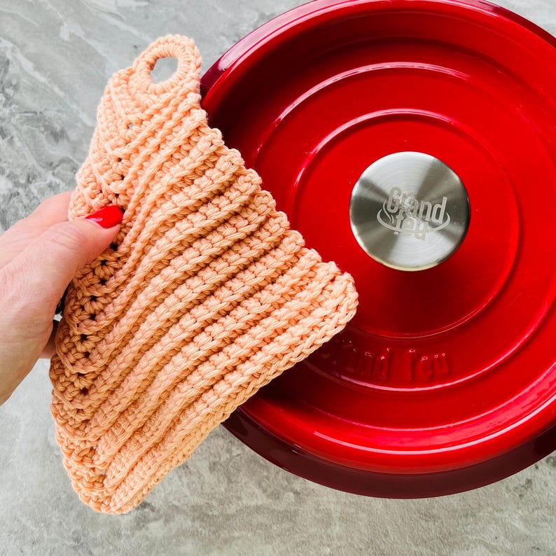 Op de afbeelding: Een perzik kleurige, gehaakte pannenlap met een lus om op te hangen. De pannenlap ligt naast een rode ge&euml;mailleerde gietijzeren pot met een zilveren handvat. De pannenlap heeft een geribbelde textuur.