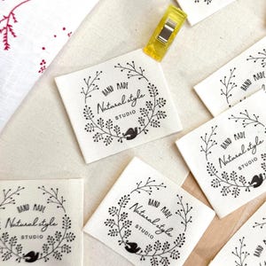 May include: A collection of square fabric labels with the text "HAND MADE Natural Style STUDIO" in black. Each label features a circular floral design with a bird silhouette. A yellow clip and a white fabric with red floral patterns are also visible.