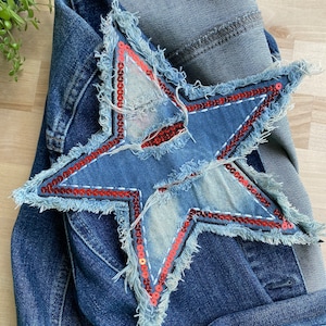 Może przedstawiać: Niebieska kurtka dżinsowa z dużą, czerwoną i niebieską naszywką w kształcie gwiazdy ozdobioną cekinami z przodu. Gwiazda ma strzępiony brzeg i jest przyszyta czerwoną nicią.