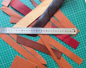 May include: Various leather strips in brown, red, and tan hues are arranged on a green cutting mat. A metal ruler, marked in centimetres, is placed across the leather. The strips have different textures and widths, indicating a craft or DIY project.