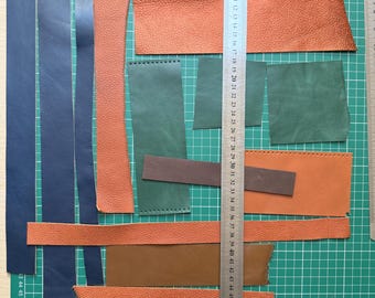 May include: A collection of leather pieces in shades of brown, orange, blue, and green, cut into different shapes. A metal ruler, marked in centimetres, is positioned across the leather. The pieces are laid out on a green cutting mat.