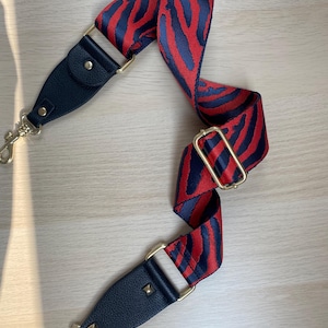 May include: A red and blue zebra print adjustable shoulder strap with gold hardware and black leather accents. The strap has two gold hooks on each end.