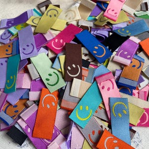 Puede incluir: Una colección de etiquetas de tela coloridas con un diseño de cara sonriente. Las etiquetas son de varios colores, incluyendo morado, rosa, azul, verde, amarillo, naranja y marrón. Las caras sonrientes están bordadas en diferentes colores de hilo.