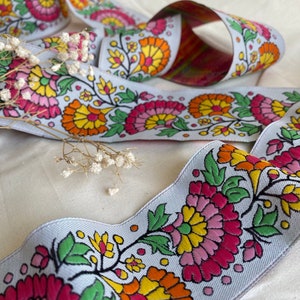 Puede incluir: Cinta de tela blanca con un estampado floral colorido. El estampado presenta flores rosas, amarillas, naranjas y verdes con contornos negros. La cinta está doblada y se muestra sobre una superficie blanca.