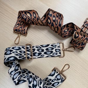 May include: Two leopard print fabric straps with gold metal clasps. One strap is brown with black spots, the other is white with black and silver glitter spots.