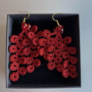 Könnte beinhalten: Ein Paar rote Häkelohrringe mit floralem Design, präsentiert in einer schwarzen Schachtel. Die Ohrringe haben goldfarbene Haken und kleine rote Perlenakzente. Die filigrane Häkelarbeit bildet ein zartes, blattartiges Muster.