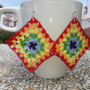 May include: Rainbow crochet earrings with a square shape and silver hooks. The earrings are hanging from a white mug.
