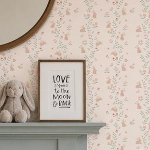 May include: A light pink wallpaper featuring a repeating pattern of small brown rabbits, green vines, and pink and orange flowers. A framed print with the text LOVE you to the MOON & BACK sits next to a plush gray rabbit on a gray mantelpiece.